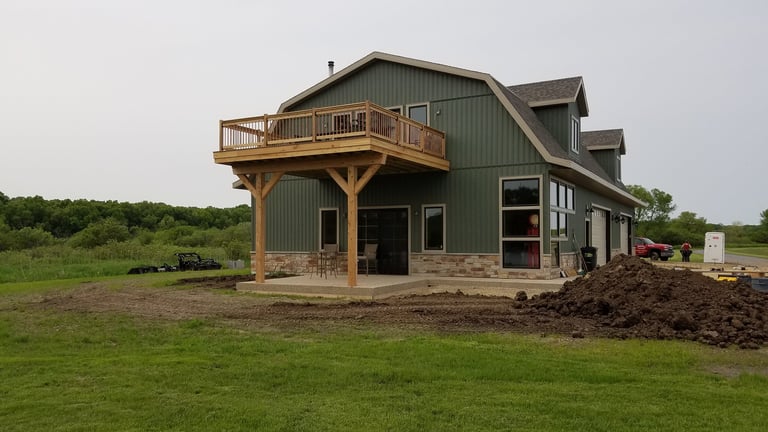 Modern barn-style house with green metal siding and wooden deck under construction in rural pasture setting