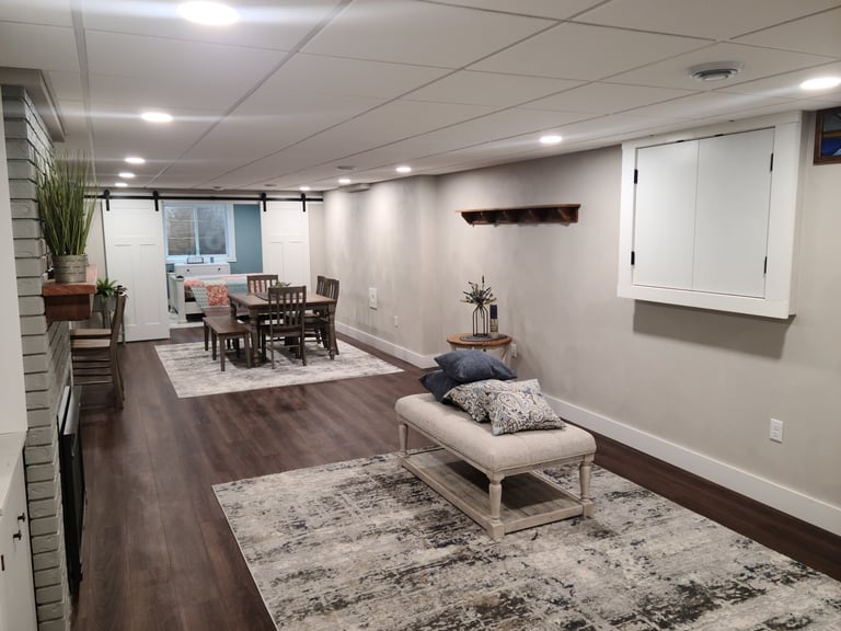 Modern basement recreation room with dining area, upholstered bench seating, area rugs, and white wall-mounted cabinets on wood flooring