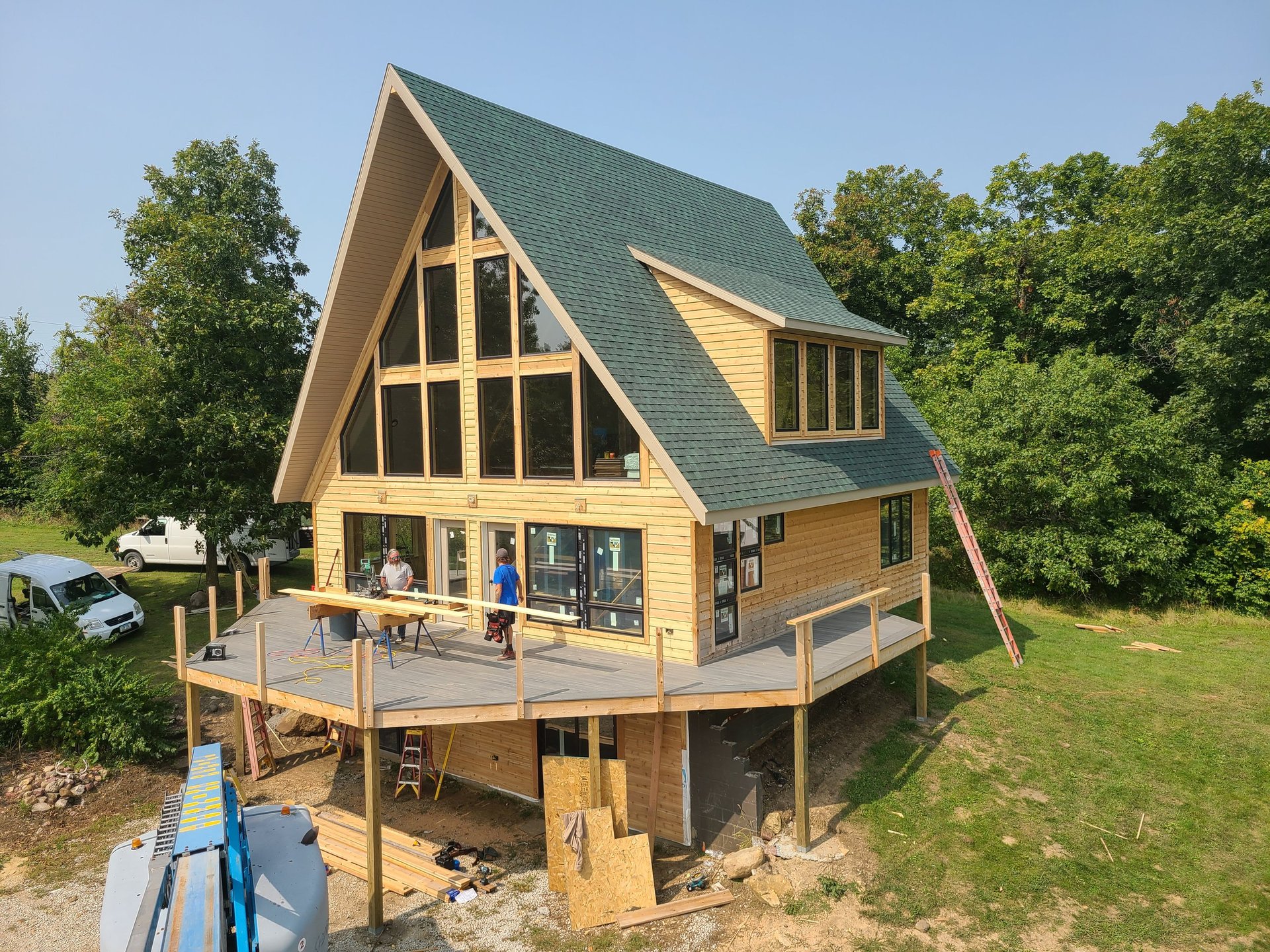 Modern A-frame house with yellow wood siding, green roof, elevated on posts with wraparound deck, surrounded by trees